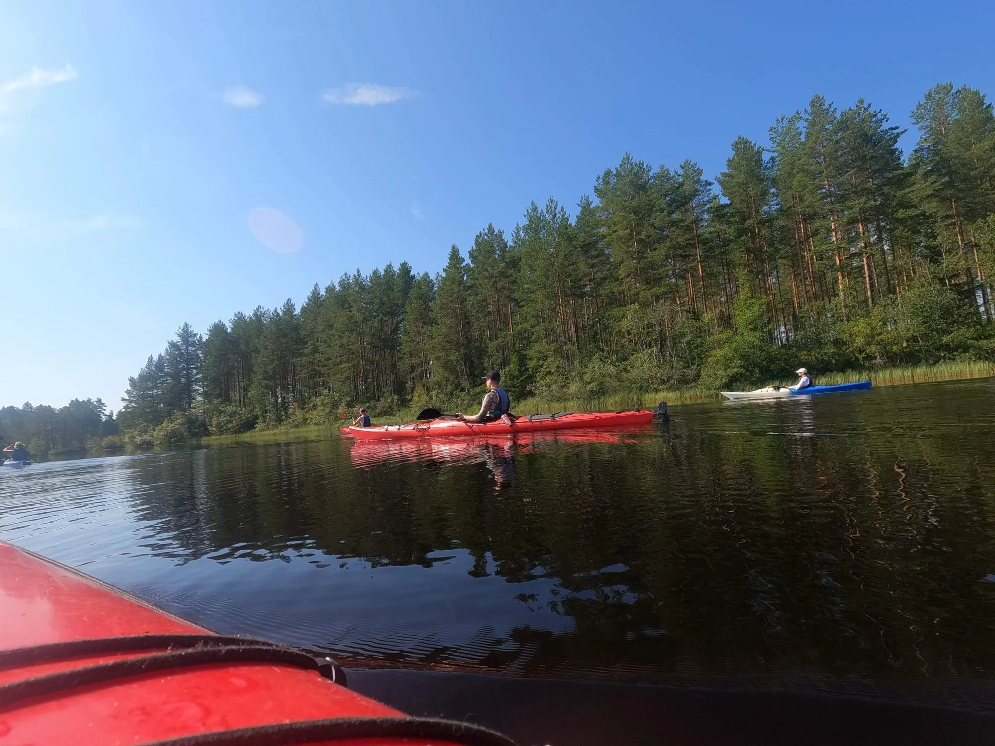 View from a red kayak on a calm lake lined with pine forest, with more kayakers ahead.